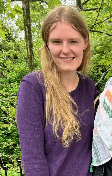 Hanna with long blond hair against green leafy backdrop wearing purple top. 