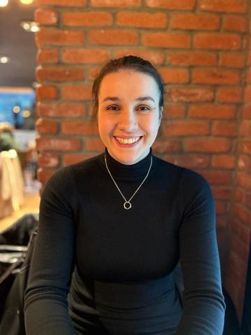Holly Woman in black wearing necklace with circle on it smiles in front of brick wall