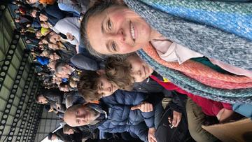Louisefootball Louise sits in the stands at a football match. She has brown hair and wearing a multicoloured scarf. Two young boys smile at the camera behind her, and so does an man behind them.