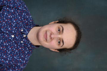 M person wearing multicoloured blue and purple dotted top with white buttons looks at camera in front of plain dark background