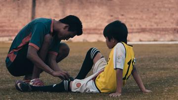 Child sits on soccer pitch while adult kneels to look at his injured leg